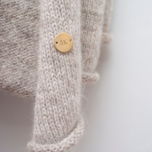 May include: A close-up of a beige knitted jumper with a gold metal tag that says "AK" on it.