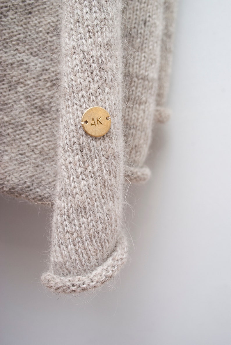 May include: A close-up of a beige knitted jumper with a gold metal tag that says "AK" on it.