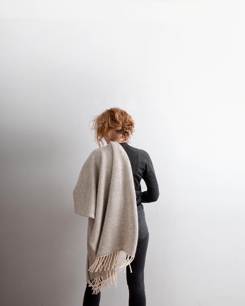 Woman from behind wearing a beige melange wool blanket scarf with off-white fringe draped over her shoulder.