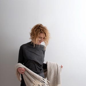 Smiling woman holding a large beige melange wool blanket scarf with off-white fringes around her waist.