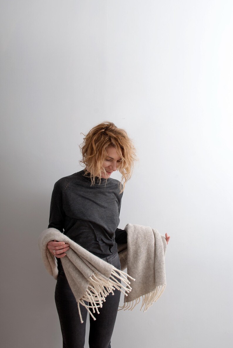 Smiling woman holding a large beige melange wool blanket scarf with off-white fringes around her waist.