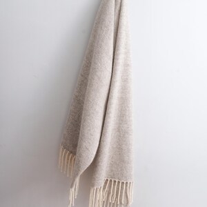 Large beige melange wool mohair blanket scarf with off-white fringe hanging gracefully on a white wall.