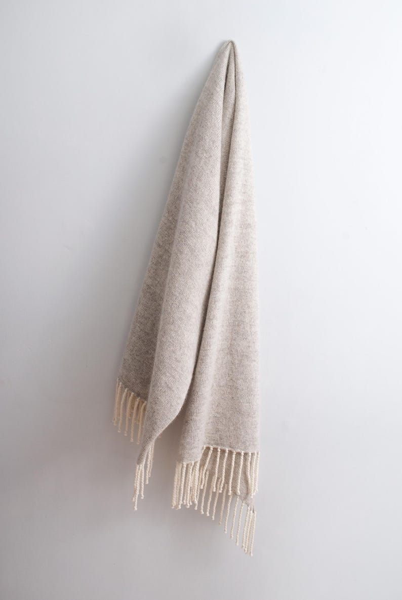 Large beige melange wool mohair blanket scarf with off-white fringe hanging gracefully on a white wall.