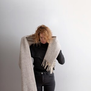 Woman wrapping a long beige melange wool blanket scarf with off-white fringe around her neck.