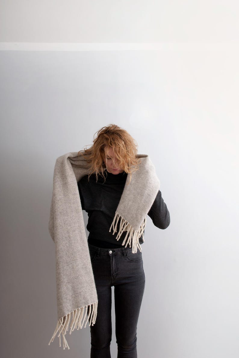 Woman wrapping a long beige melange wool blanket scarf with off-white fringe around her neck.