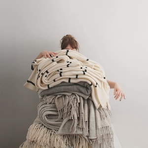 A stack of blankets in various shades of white and gray, some with fringe, are piled high. Textile artist Agnese is partially hidden behind the blankets.