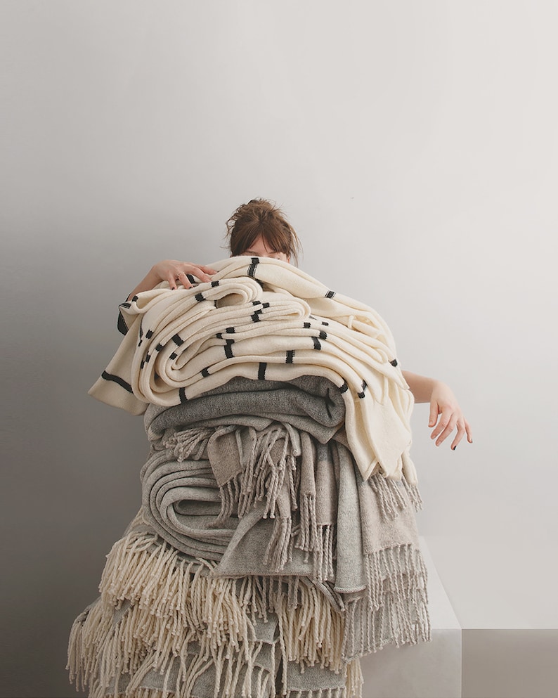 A stack of blankets in various shades of white and gray, some with fringe, are piled high. Textile artist Agnese is partially hidden behind the blankets.