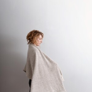 Side profile view of a woman walking, wearing a beige wool blanket scarf with off-white fringe. Large, thick and cozy feelings.