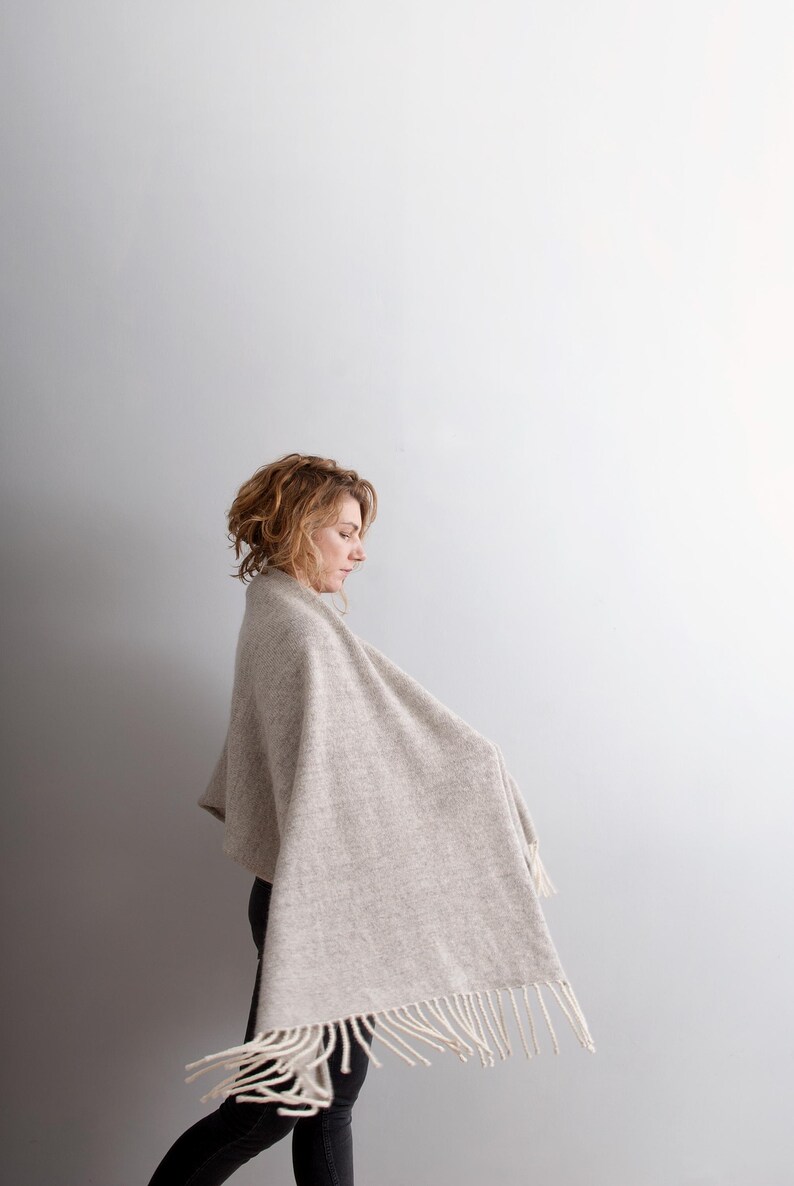 Side profile view of a woman walking, wearing a beige wool blanket scarf with off-white fringe. Large, thick and cozy feelings.