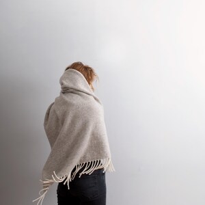 Back view of a woman wearing a thick beige wool blanket shawl wrapped over her head and shoulders.
