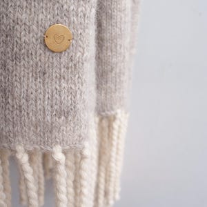 Close-up detail of a beige melange wool blanket scarf with off-white fringe and a brass heart pin.
