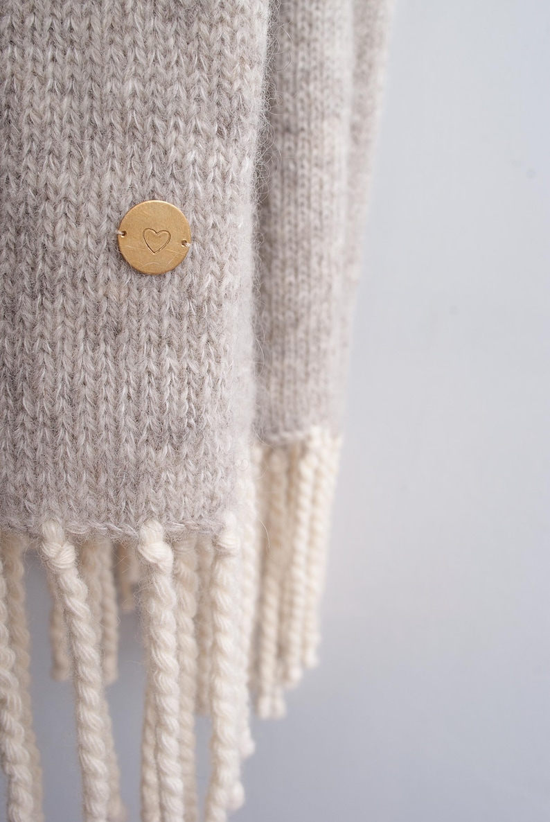Close-up detail of a beige melange wool blanket scarf with off-white fringe and a brass heart pin.