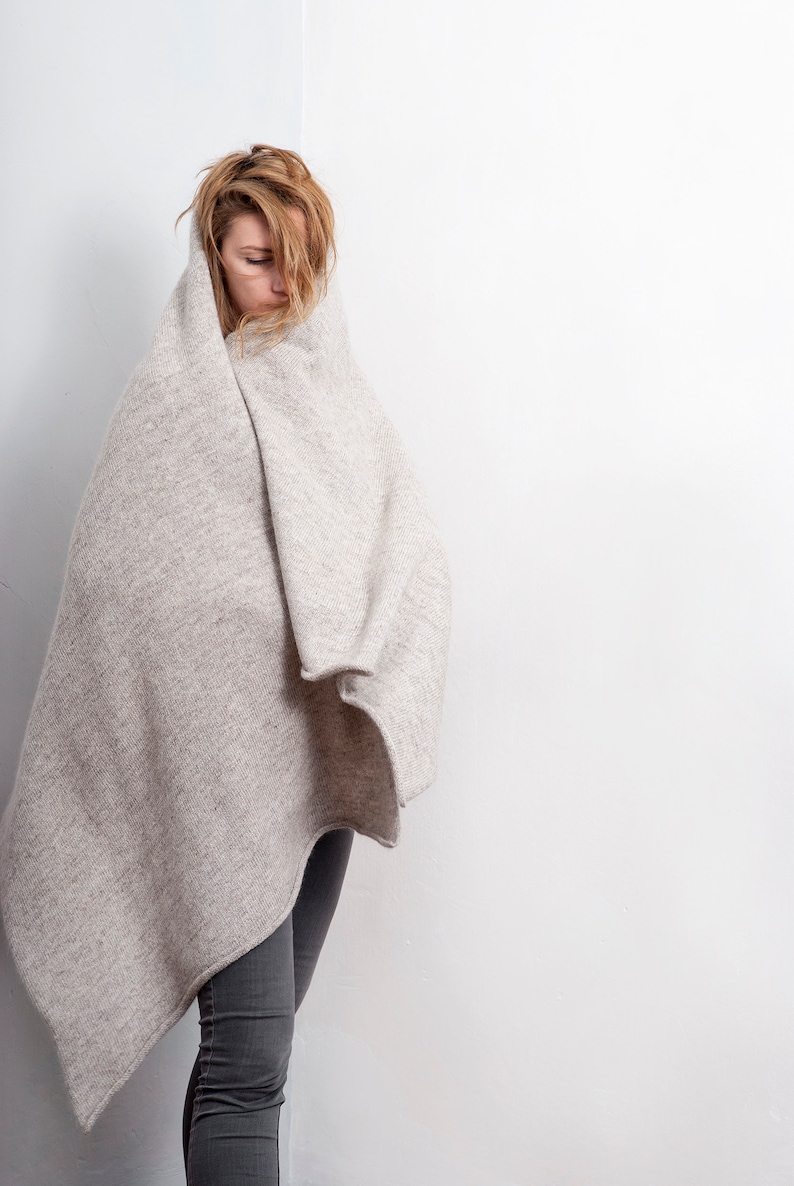 May include: A woman wearing grey jeans is wrapped in a large, light grey blanket. She is looking down and her hair is covering her face.