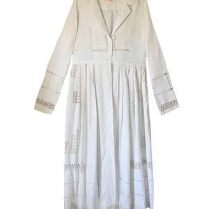 May include: A white long-sleeved dress with a button-up front and intricate lace detailing throughout. The dress has a fitted bodice and a flowing skirt.