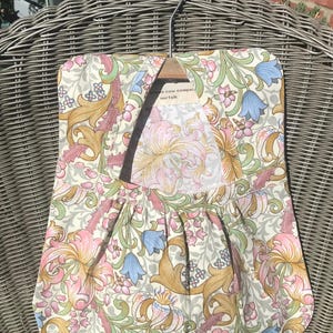 May include: A floral patterned apron with a cream background, featuring pink, blue, and gold flowers and green leaves. The apron is hanging on a wooden hanger. The text on the tag reads "The cow company, norfolk."