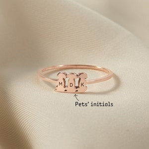May include: A rose gold ring with three dog silhouettes, each with an initial engraved inside. The initials are M, D, and K. The ring is on a light brown fabric background.