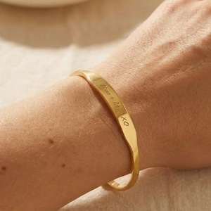 Peut inclure: Un bracelet manchette en métal doré à la surface lisse et légèrement incurvée. Le bracelet présente une inscription gravée qui se lit "Mum + Dad XO" en écriture cursive, positionnée le long du centre de la bande sur le poignet.