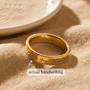 Könnte beinhalten: Ein goldfarbener Metallring mit individueller Gravur auf der Außen- und Innenseite. Das äußere Band zeigt den kursiven Text "Love you Hanna" mit einem Pfeil, der auf den Schriftzug "actual handwriting" zeigt.