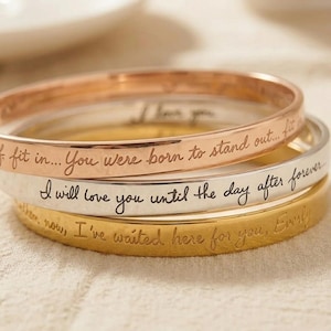 Könnte beinhalten: Ein Stapel aus drei Metallarmreifen in Roségold-, Silber- und Goldtönen. Jeder Armreif ist mit einer kursiven Gravur versehen. Das roségoldene Band trägt den Text "You were born to stand out... fit in...", das silberne Band "I will love you until the day after forever" und das goldene Band "I've waited here for you, Everly".