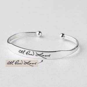 May include: A silver-toned open cuff bracelet featuring a polished finish and rounded ball ends. The front surface of the bracelet is engraved with the cursive black text "All Our Love," which matches the handwritten sample shown below the bracelet.