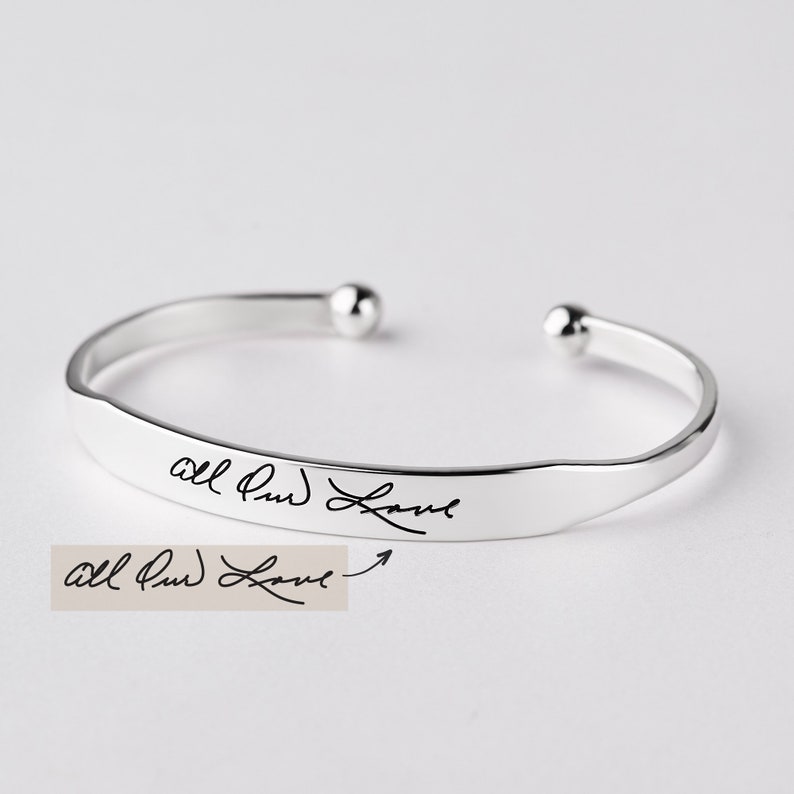 May include: A silver-toned open cuff bracelet featuring a polished finish and rounded ball ends. The front surface of the bracelet is engraved with the cursive black text "All Our Love," which matches the handwritten sample shown below the bracelet.