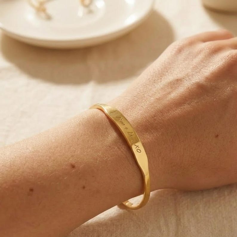 May include: A gold-toned metal cuff bracelet with a smooth, slightly curved surface. The bracelet features an engraved inscription that reads "Mum + Dad XO" in a cursive script, positioned along the center of the band on the wrist.