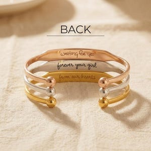May include: A set of three open cuff bracelets in rose gold, silver, and gold tones. Each bracelet features a spherical end and an engraved inscription. The top reads 'waiting for you', the middle 'forever your girl', and the bottom 'from our hearts'.
