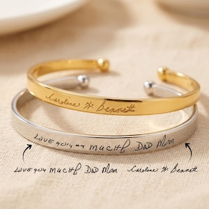May include: Two open cuff bracelets, one gold-toned and one silver-toned, each featuring engraved handwriting. The gold bracelet is inscribed with the name Caroline H. Bennett, while the silver bracelet displays the handwritten text Love you much, Dad Mom.