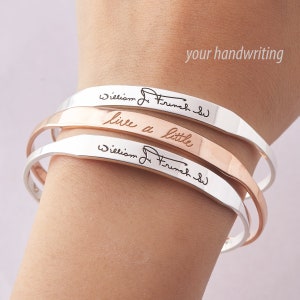 May include: Three metal cuff bracelets stacked together. Two silver-toned cuffs feature the black engraved signature William J. French Sr. The middle rose gold-toned cuff is engraved with the cursive text live a little. The text your handwriting is visible.