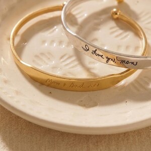 May include: Two open cuff bracelets, one gold-toned and one silver-toned. The gold bracelet is engraved with the text "Mom & Dad XO". The silver bracelet features the black engraved script text "I love you, mom" and has rounded ball ends.