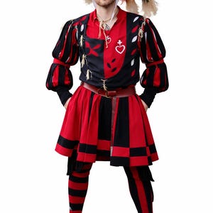Op de afbeelding: Een persoon in een Renaissance-kostuum. De outfit heeft een zwart en rood kleurenschema, inclusief een hoed met veren, een patroonshirt, een rok en gestreepte leggings. Een bruine riem en schoenen maken de outfit compleet.