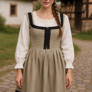 Pode incluir: Uma mulher vestindo um vestido tradicional. O vestido verde-oliva tem acabamentos pretos e detalhes florais brancos. O vestido é usado sobre uma blusa branca com mangas compridas e decote com babados.
