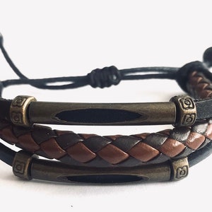 Pode incluir: Um bracelete de couro trançado com dois detalhes metálicos. O bracelete é castanho e preto e tem um fecho de cordão preto.