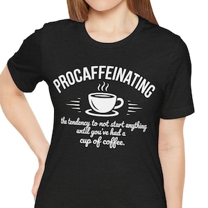 May include: Black t-shirt with the word "PROCAFFEINATING" above a coffee cup graphic. Below the graphic, the text reads "the tendency to not start anything until you've had a cup of coffee."