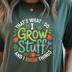 May include: Dark green t-shirt with the text "THAT'S WHAT I DO I GROW STUFF AND I KNOW THINGS" in a circular design. The design includes gardening tools and foliage in green, orange, and blue. The shirt is a relaxed fit.