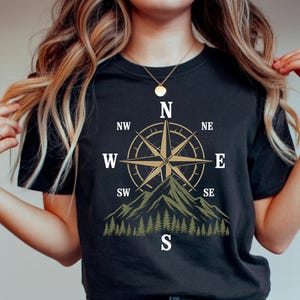 May include: A black short-sleeve t-shirt featuring a graphic print of a gold compass rose centered above a green mountain range and a forest of pine trees. The compass includes the cardinal and intercardinal directions labeled N, NE, E, SE, S, SW, W, and NW in white text.