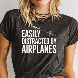 May include: Dark gray t-shirt with the text "EASILY DISTRACTED BY AIRPLANES" in white. The design includes a small airplane and a starburst graphic. The shirt is a crew neck style.