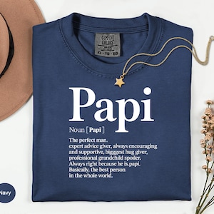 May include: Navy blue t-shirt with the word "Papi" in large white letters. The shirt includes a definition of "Papi" below the word. A gold star pendant hangs from a gold chain. The shirt is on a white surface.