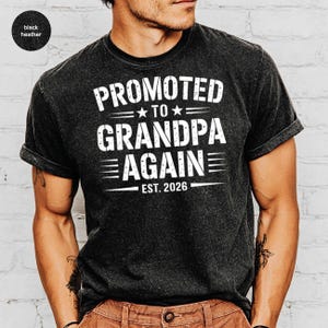 May include: Black heather t-shirt with the text "PROMOTED TO GRANDPA AGAIN EST. 2026" in white. The shirt has a crew neck and short sleeves. The text is centered on the front of the shirt.