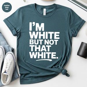 May include: Heather deep teal t-shirt with the text "I'M WHITE BUT NOT THAT WHITE" in white block letters. The shirt is knotted at the bottom and is paired with white sneakers and blue jeans.