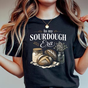May include: Black t-shirt with the text "In my Sourdough Era" and illustrations of bread, jars, and wheat. The design features a rustic aesthetic, perfect for bakers and bread enthusiasts.