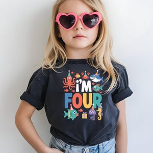 May include: A young person wearing a black t-shirt with the words "I'm FOUR" in colorful letters and sea creature graphics. The person is also wearing pink heart-shaped sunglasses and blue jeans.