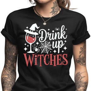 May include: Black t-shirt with a Halloween-themed graphic. The design features a witch's hat, a wine glass filled with red liquid, a spider web, and the words "Drink up WITCHES" in white and red.