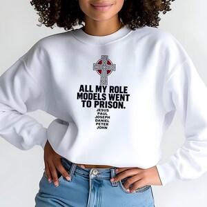 May include: A white long-sleeve crewneck sweatshirt featuring a graphic of a Celtic cross above the text "ALL MY ROLE MODELS WENT TO PRISON." followed by a list of names: "JESUS, PAUL, JOSEPH, DANIEL, PETER, JOHN" printed in black ink on the chest.