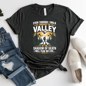 May include: Dark gray t-shirt with a graphic featuring a hiker in a valley scene, with text that reads "EVEN THOUGH I WALK THROUGH THE VALLEY OF THE SHADOW OF DEATH I WILL FEAR NO EVIL..." Psalm 23:4.