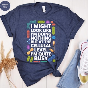 May include: Heather navy blue t-shirt with the text "I might look like I'm doing nothing but at the cellular level I'm quite busy" and colorful science-themed graphics. The shirt is paired with blue jeans and white sneakers.