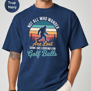 May include: True navy blue t-shirt with a Bigfoot graphic and the text "NOT ALL WHO WANDER ARE LOST SOME ARE LOOKING FOR Golf Balls". The graphic features a Bigfoot silhouette walking in front of a sunset.