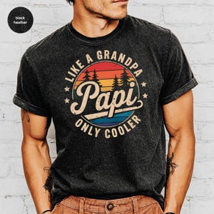 May include: Black heather t-shirt with a circular graphic that reads "LIKE A GRANDPA PAPI ONLY COOLER" in a vintage style. The graphic features a sunset design with trees and stars. The shirt has rolled-up sleeves.