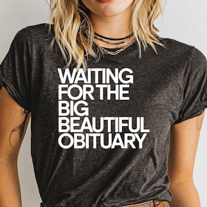 May include: Dark gray t-shirt with the text "WAITING FOR THE BIG BEAUTIFUL OBITUARY" in white block letters. The shirt has short sleeves and a crew neck.