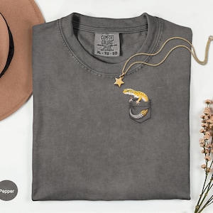 May include: Pepper-colored t-shirt with a pocket featuring a yellow and black gecko design. A gold necklace with a star pendant rests on the shirt. The shirt is a Comfort Colors XL size.